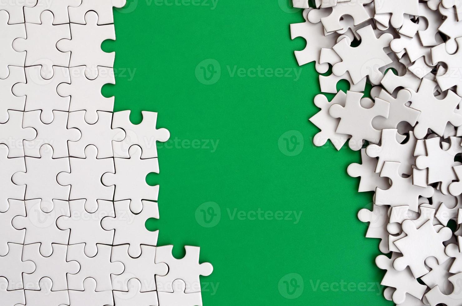 Fragment of a folded white jigsaw puzzle and a pile of uncombed puzzle elements against the background of a green surface. Texture photo with space for text