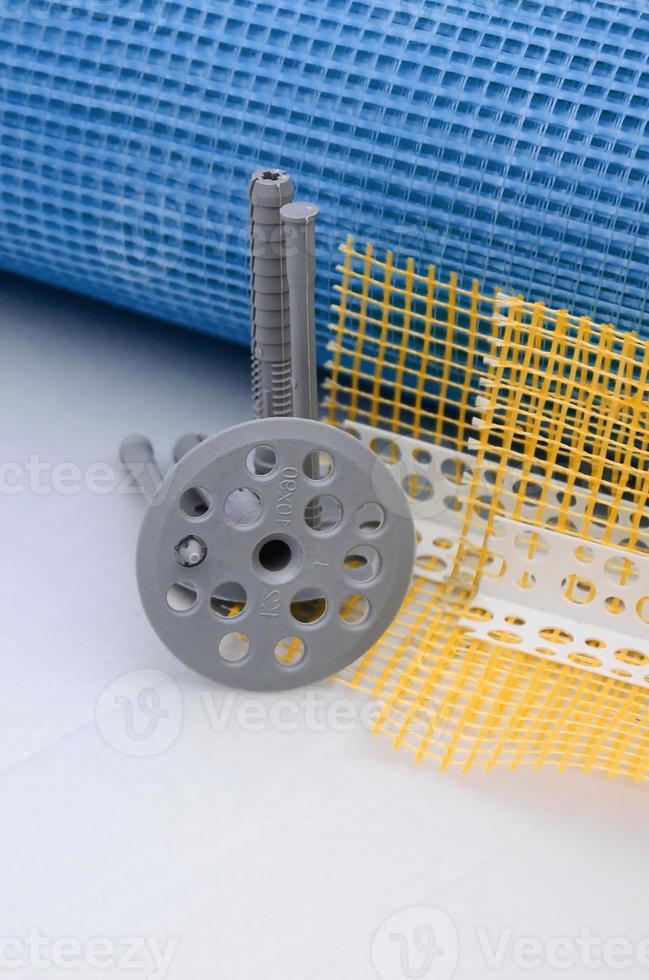 A set of construction items for the insulation of walls. Plastic dowels, a roll of mesh for the insulation of facades and a corner profile with a grid lie on the foam polystyrene surface photo
