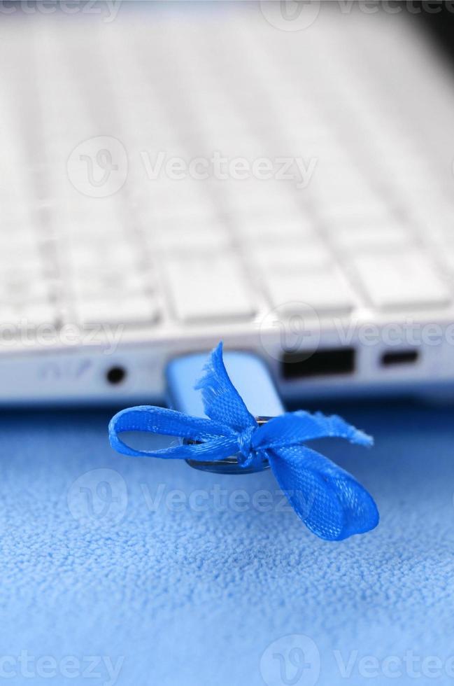 A brilliant blue USB flash drive with a blue bow is connected to a white laptop, which lies on a blanket of soft and fluffy light blue fleece fabric. Classic female design for a memory card photo