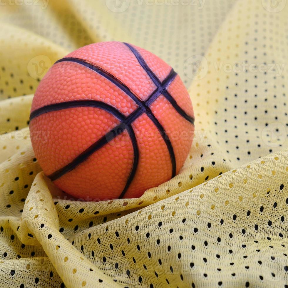 Small orange rubber basketball lies on a yellow sport jersey clothing fabric texture and background with many folds photo