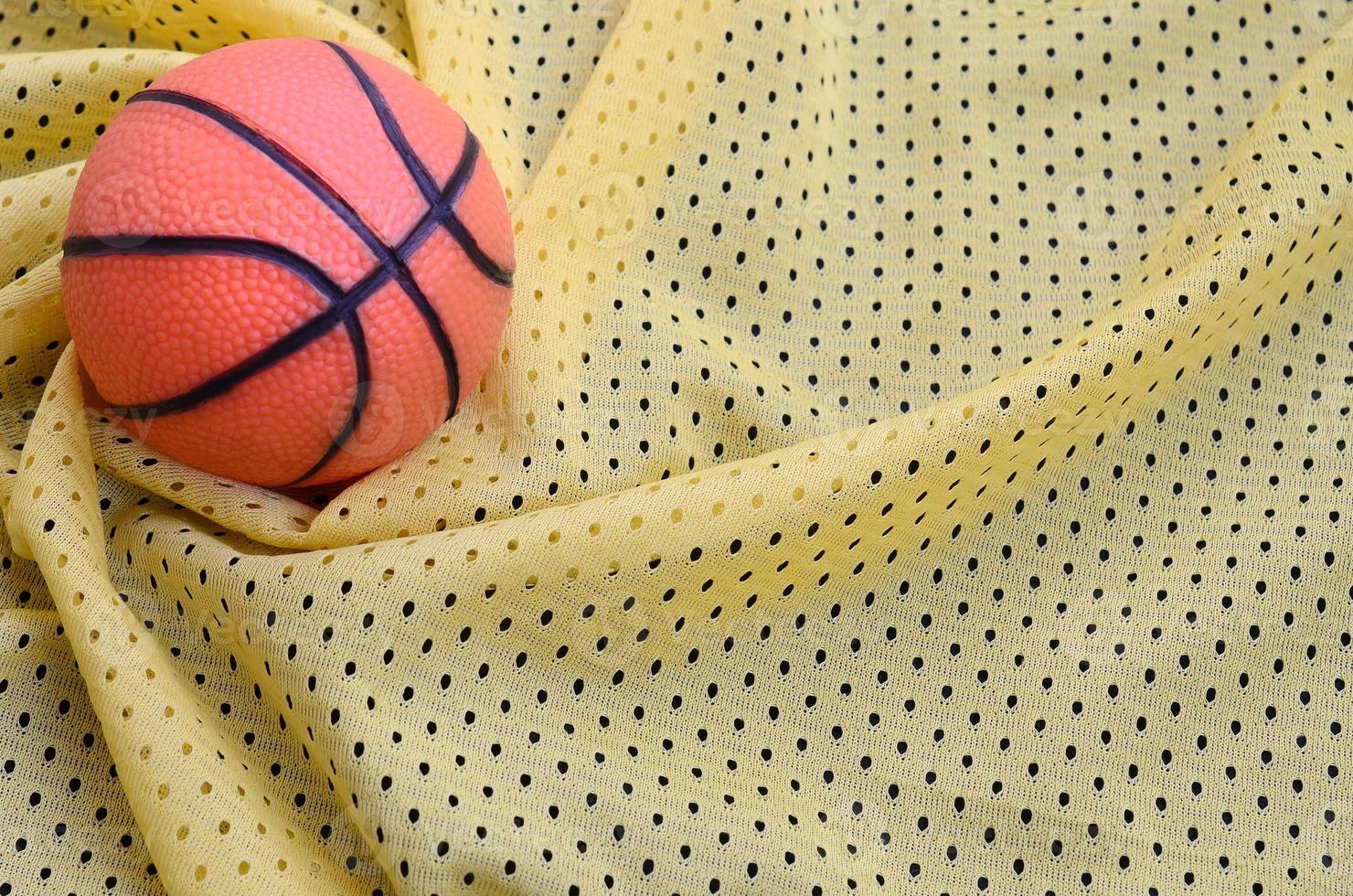 el pequeño baloncesto de goma naranja se encuentra sobre una textura de tela de jersey deportivo amarillo y un fondo con muchos pliegues foto