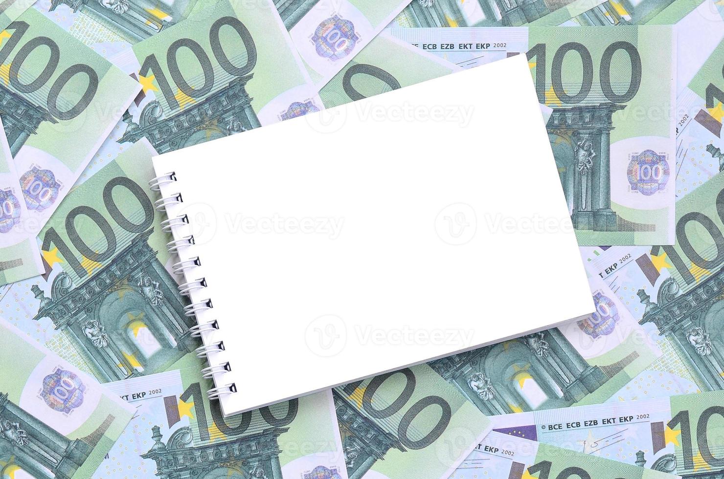 White notebook with clean pages lying on a set of green monetary denominations of 100 euros. A lot of money forms an infinite heap photo