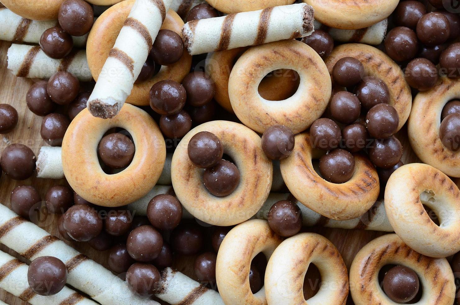 A lot of crispy sweet tubules, chocolate melting balls and yellow bagels lie on a wooden surface. Close up view photo