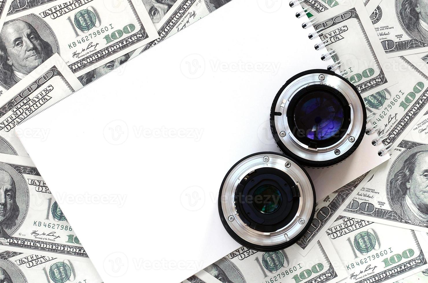 Two photographic lenses and white notebook lie on the background of a lot of dollar bills. Space for text photo