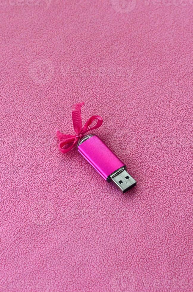 la tarjeta de memoria flash usb de color rosa brillante con un lazo rosa yace sobre una manta de suave y peluda tela de vellón rosa claro. diseño clásico de regalo femenino para una tarjeta de memoria foto