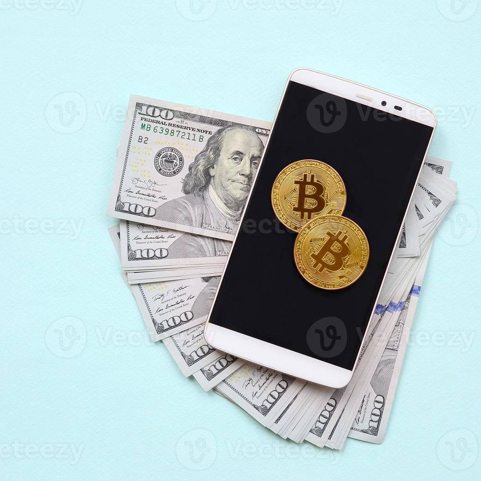 Bitcoins lies on a smartphone and hundred dollar bills on a light blue background photo