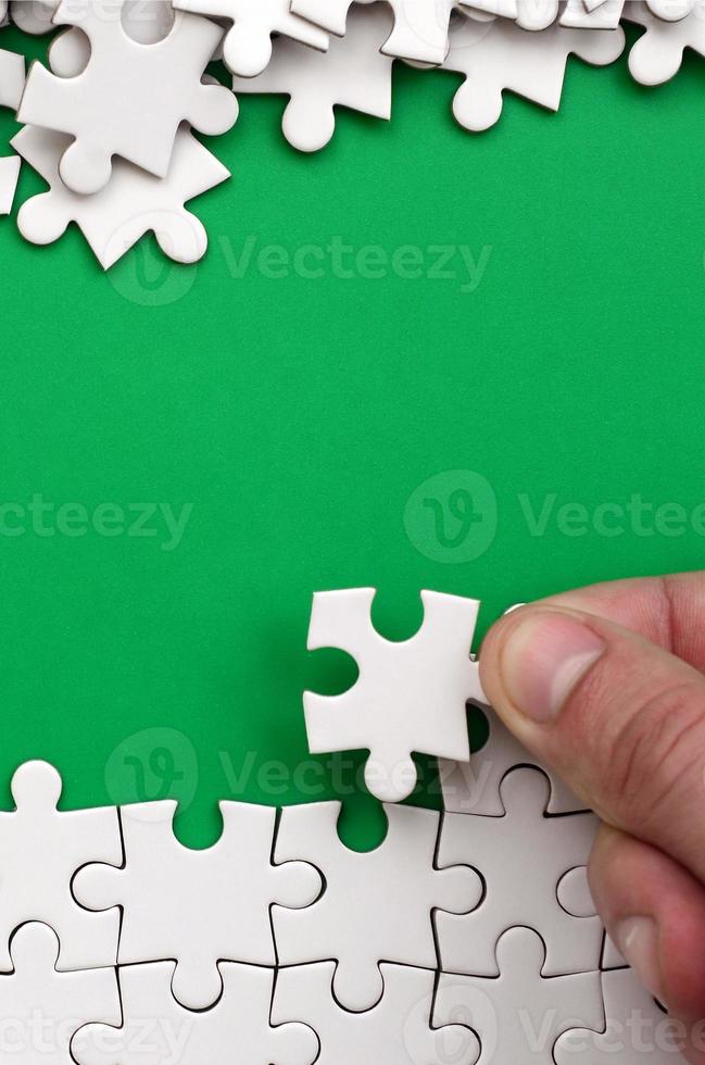 The hand folds a white jigsaw puzzle and a pile of uncombed puzzle pieces lies against the background of the green surface. Texture photo with space for text