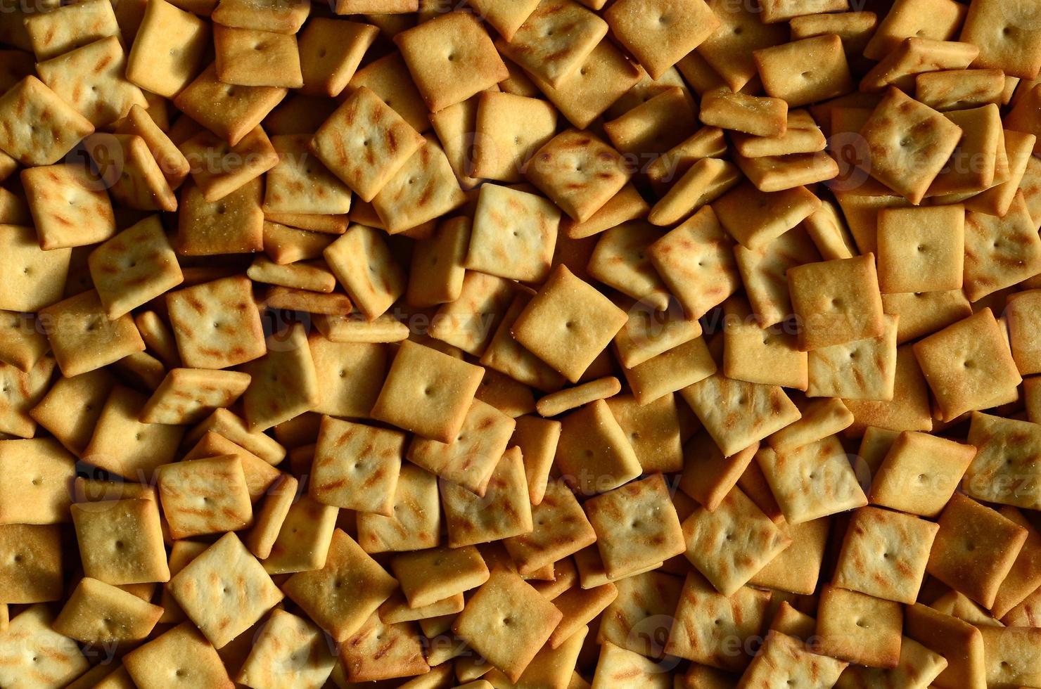 A lot of small cookies are square shaped. A pattern of a yellow salt cracker. Background image with salted pastry photo