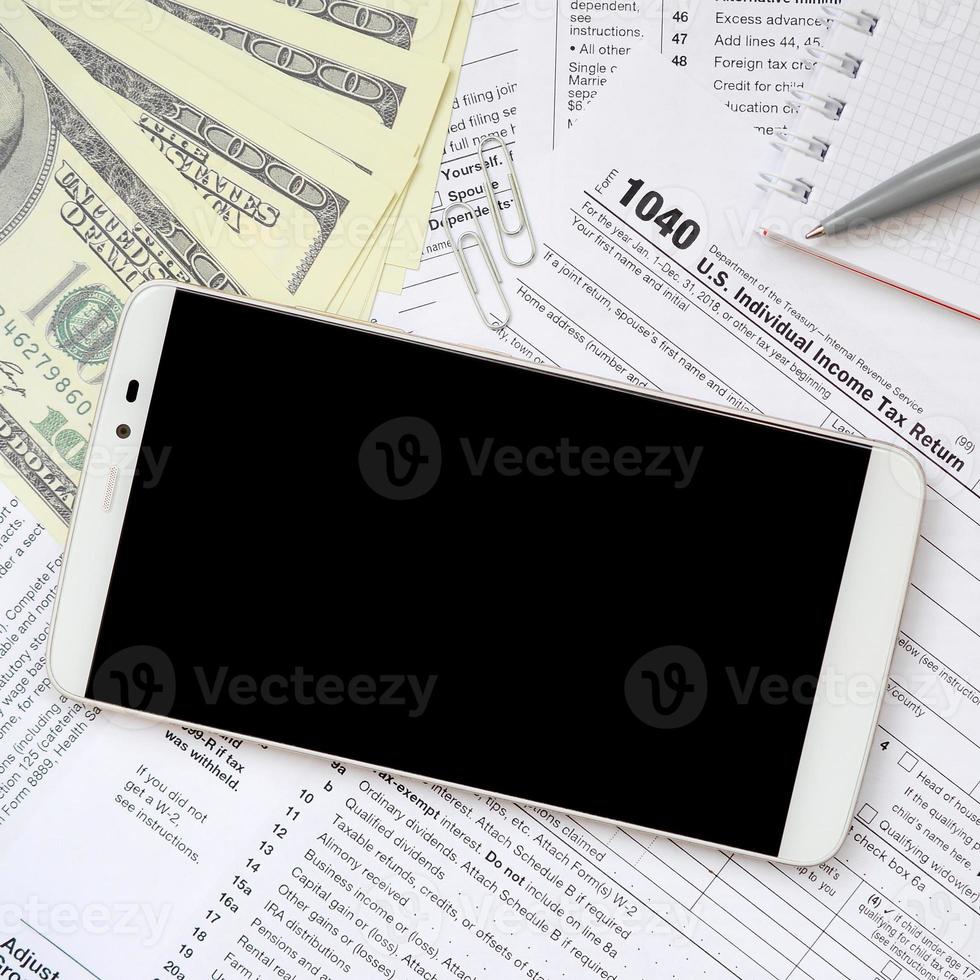 Composition of items lying on the 1040 tax form. Dollar bills, pen, smartphone, paper clip and notepad. Clean black screen smartphone for text placement photo