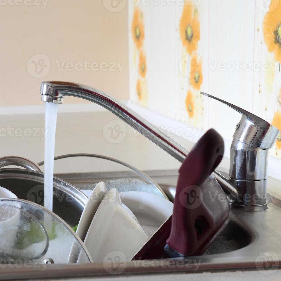 Dirty dishes and unwashed kitchen appliances lie in foam water under a tap from a kitchen faucet photo