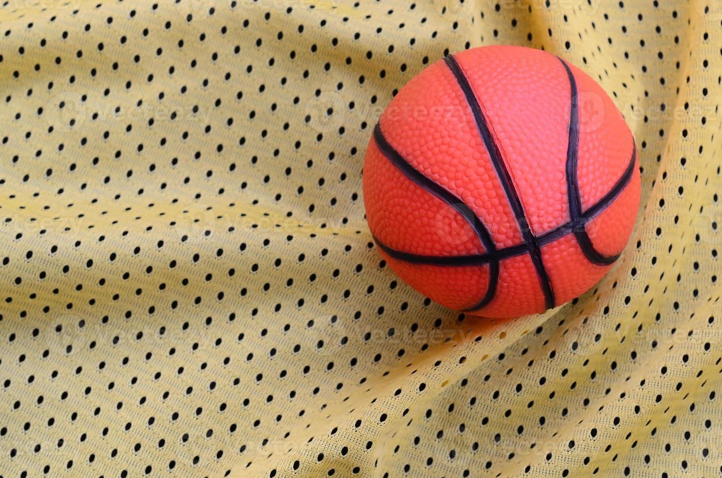 Small orange rubber basketball lies on a yellow sport jersey clothing fabric texture and background with many folds photo