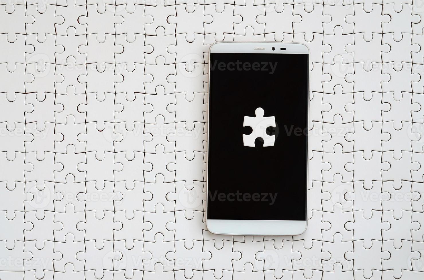 A modern big smartphone with several puzzle elements on the touch screen lies on a white jigsaw puzzle in an assembled state photo