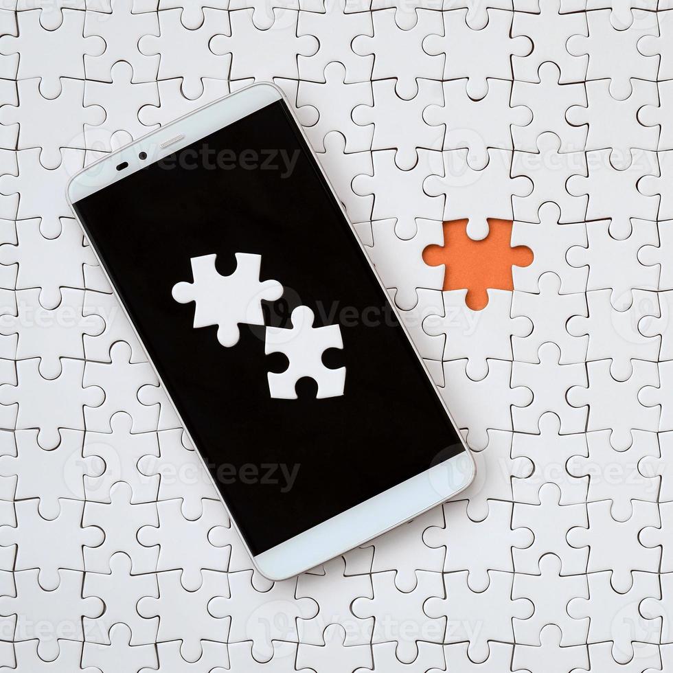 A modern big smartphone with several puzzle elements on the touch screen lies on a white jigsaw puzzle in an assembled state with missing elements photo