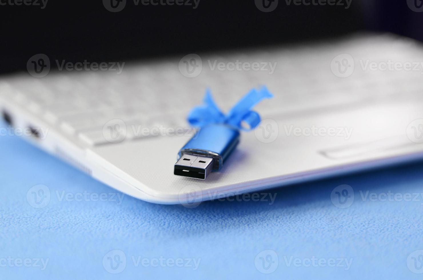 Brilliant blue usb flash memory card with a blue bow lies on a blanket of soft and furry light blue fleece fabric beside to a white laptop. Classic female gift design for a memory card photo