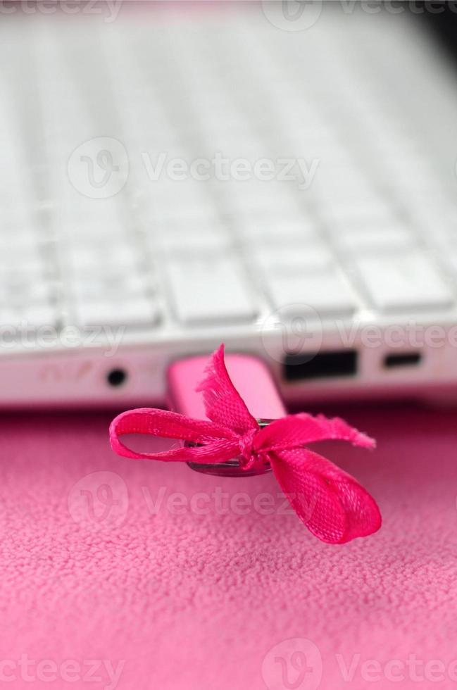 A brilliant pink USB flash drive with a pink bow is connected to a white laptop, which lies on a blanket of soft and fluffy light pink fleece fabric. Classic female design for a memory card photo