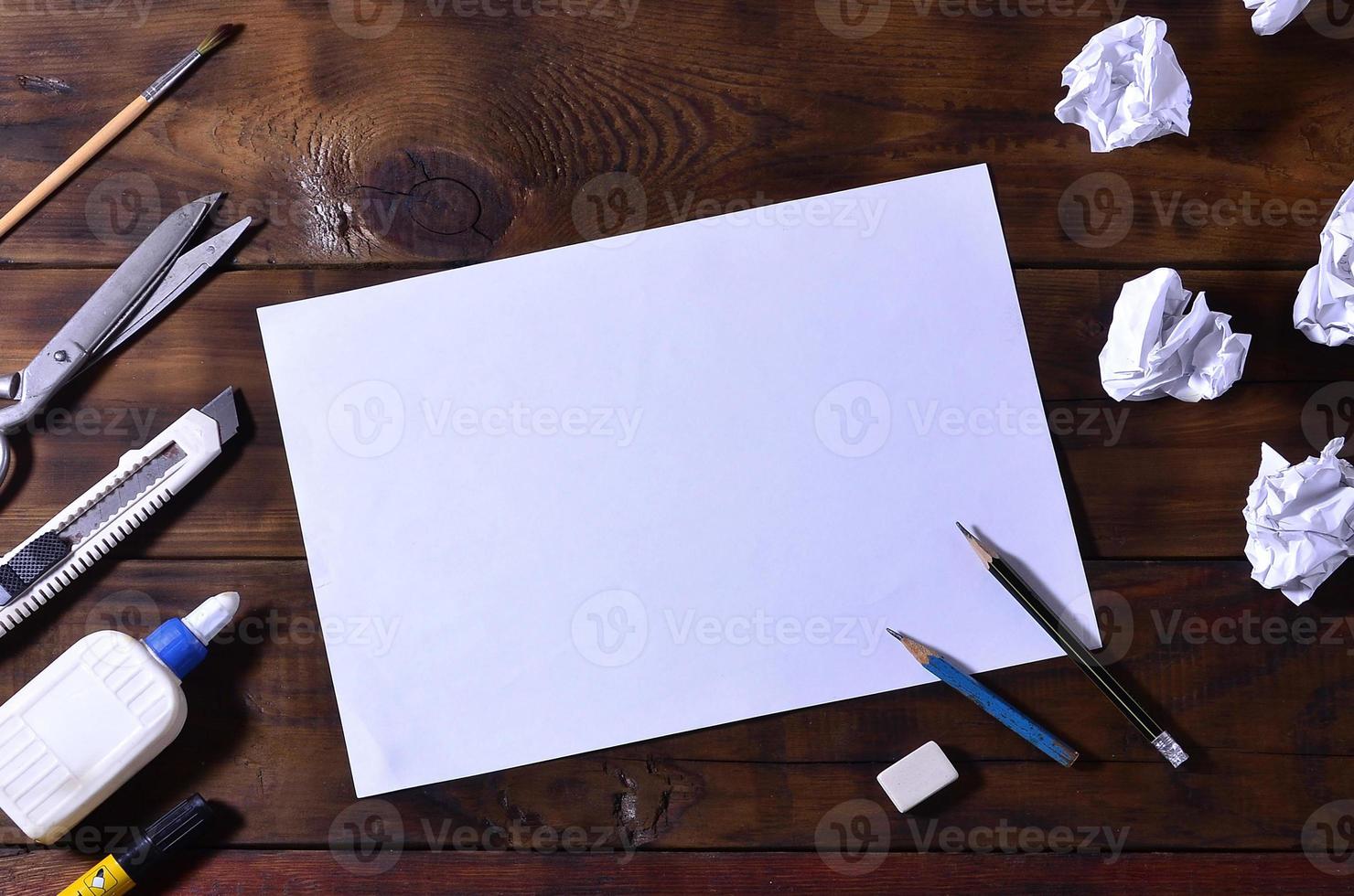 A school or office still life with a white blank sheet of paper and many office supplies. The school supplies lie on a brown wooden background. Place for text photo