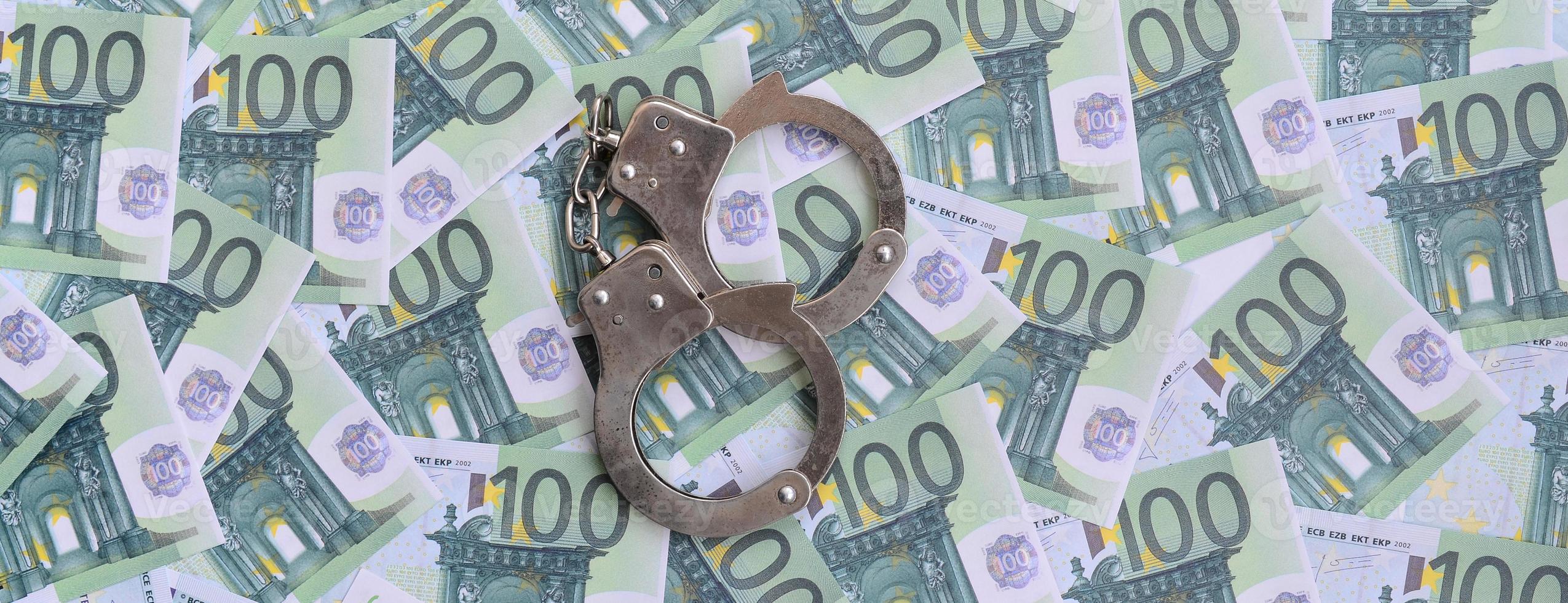 Police handcuffs lies on a set of green monetary denominations of 100 euros. A lot of money forms an infinite heap photo