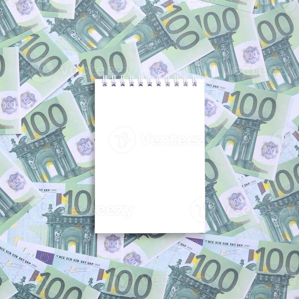 cuaderno blanco con páginas limpias sobre un conjunto de denominaciones monetarias verdes de 100 euros. mucho dinero forma un montón infinito foto