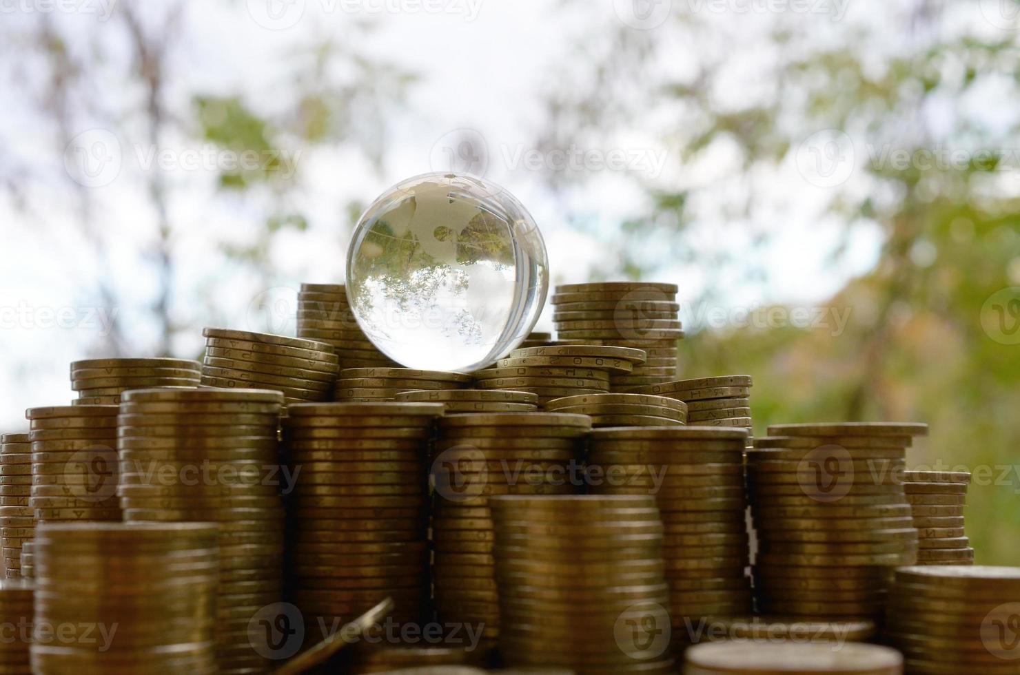 Glass planet on big pile of shiny ukrainian old 1 hryvnia coin stacks close up on blurred green trees background. The concept of money transfer around whole world photo