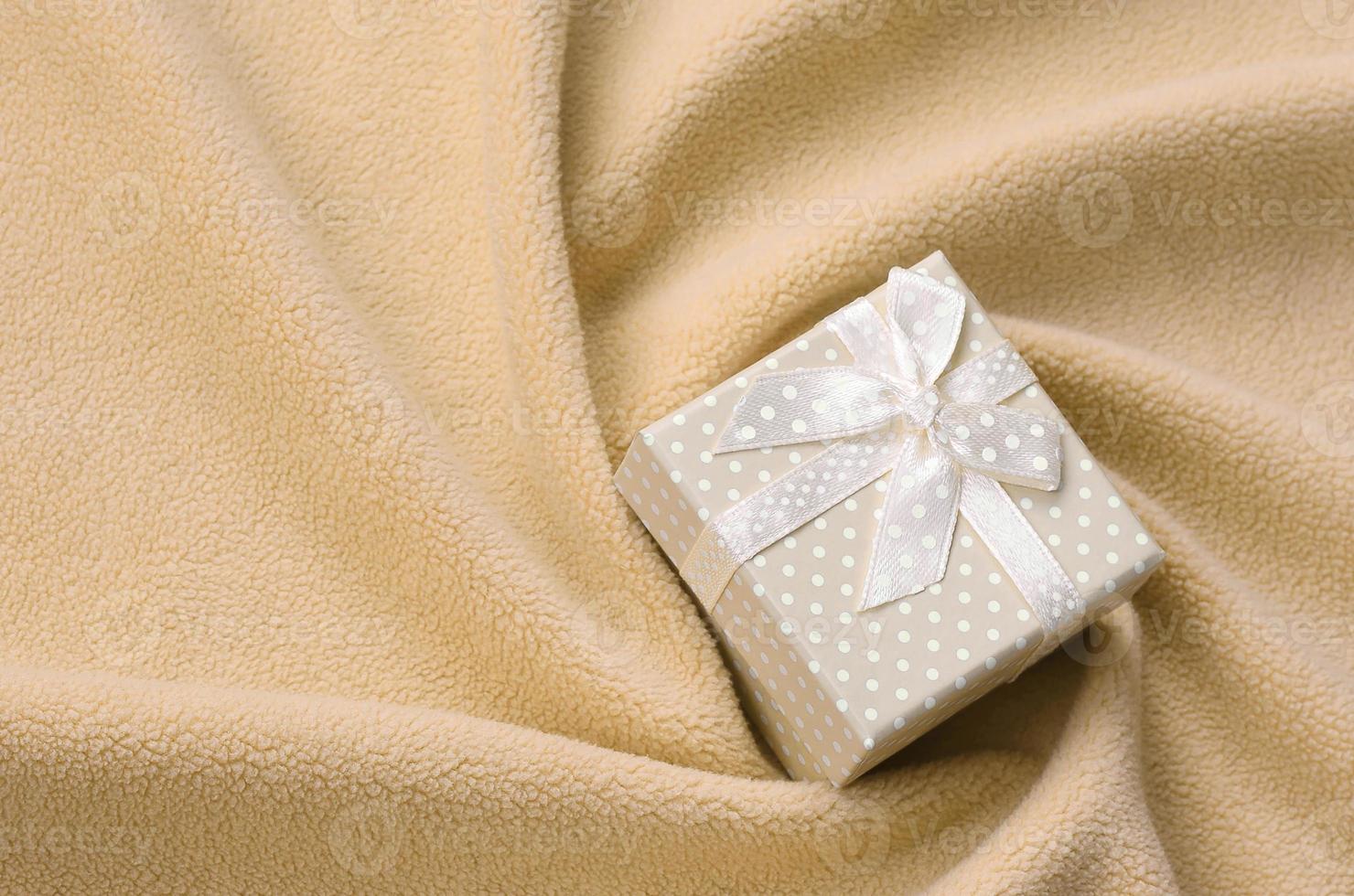 A small gift box in orange with a small bow lies on a blanket of soft and furry light orange fleece fabric with a lot of relief folds. Packing for a gift to your lovely girlfriend photo