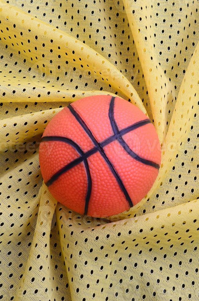 Small orange rubber basketball lies on a yellow sport jersey clothing fabric texture and background with many folds photo