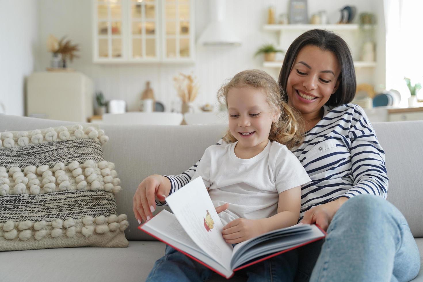 feliz madre adoptiva, hija adoptiva preescolar leyendo un libro de cuentos de hadas, sentada en el sofá en casa foto