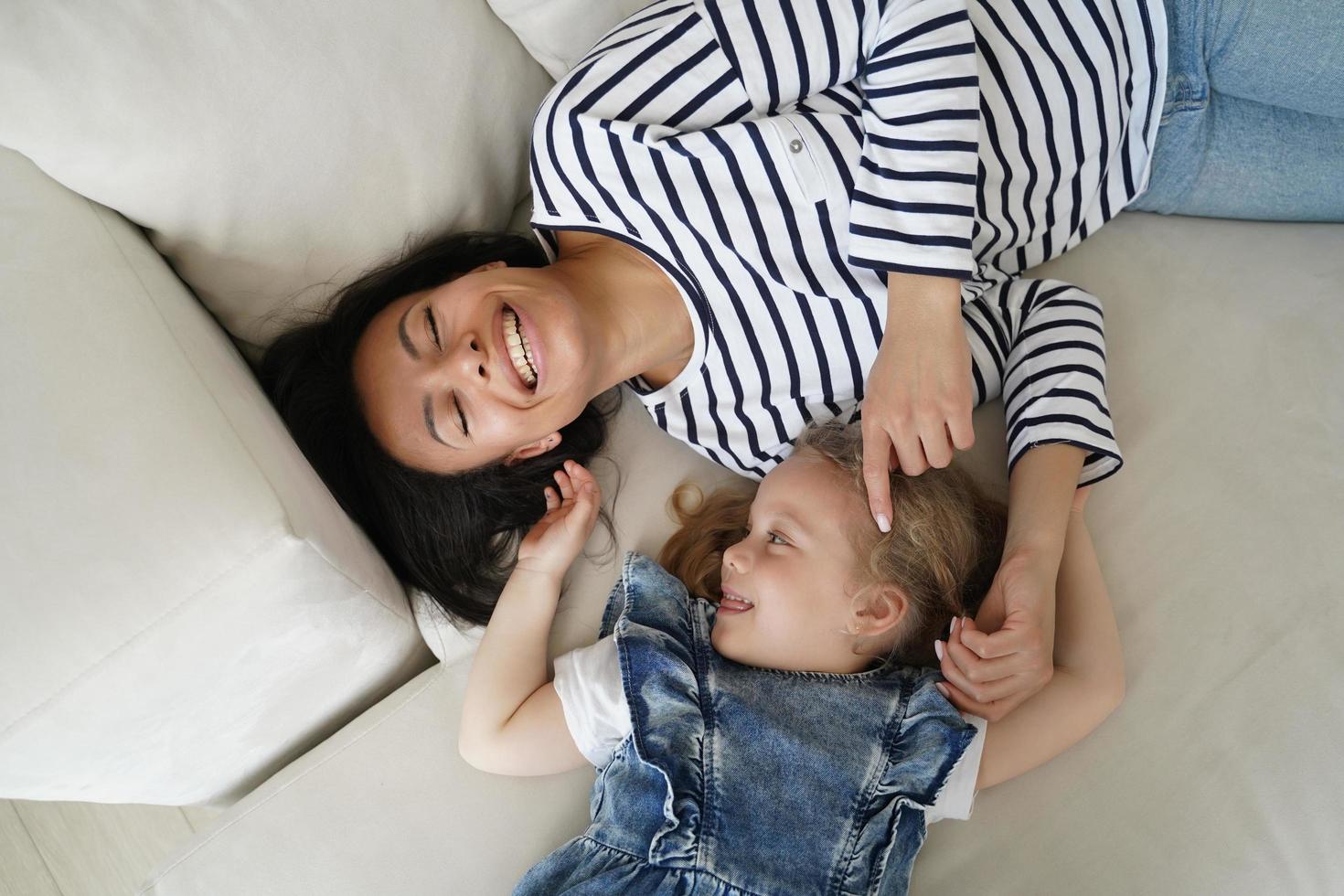 Happy adopted little kid girl tickling laughing mother, lying, having fun together on sofa, top view photo