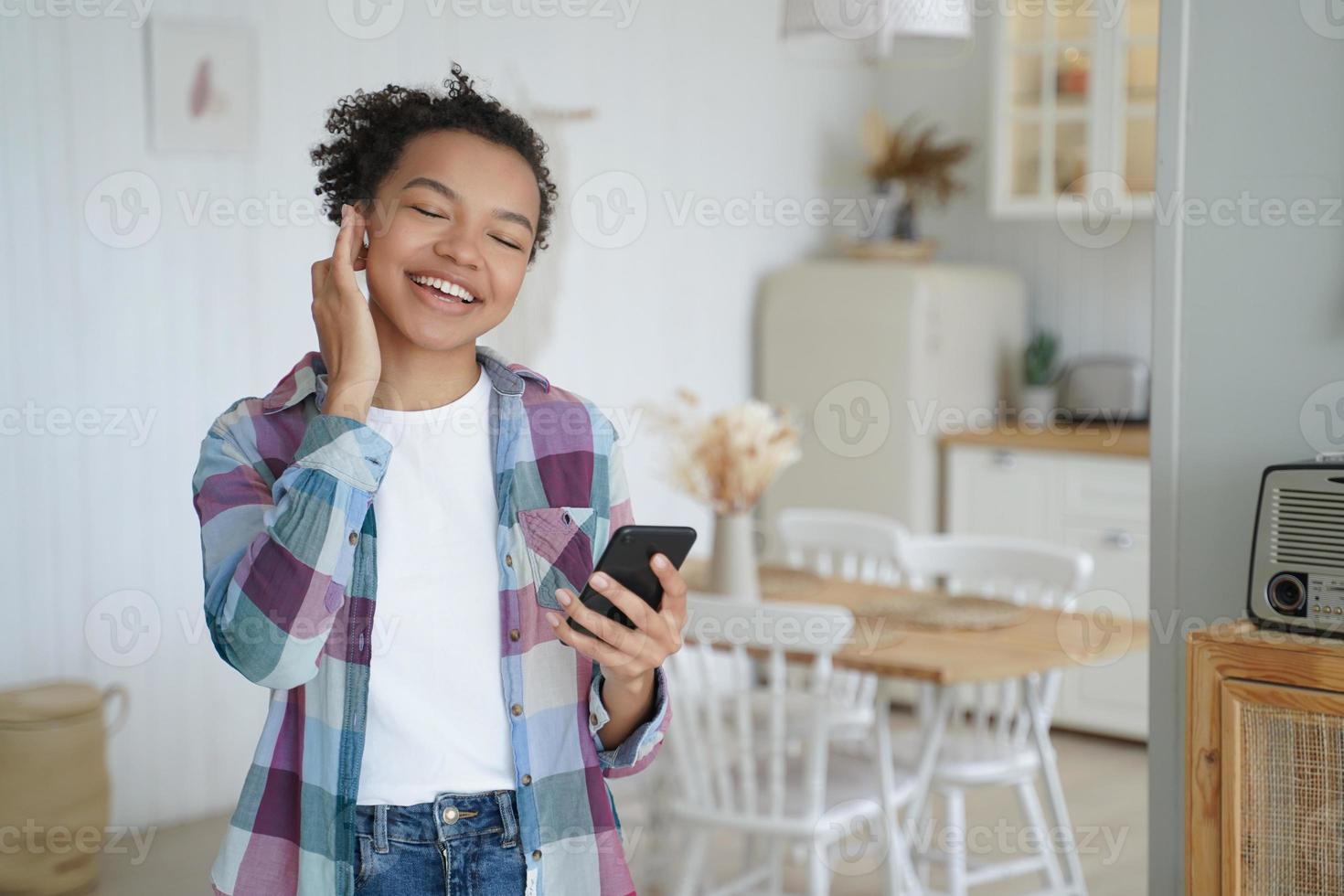 Happy mixed race girl in earphone holds smartphone using modern mobile apps, listening music at home photo