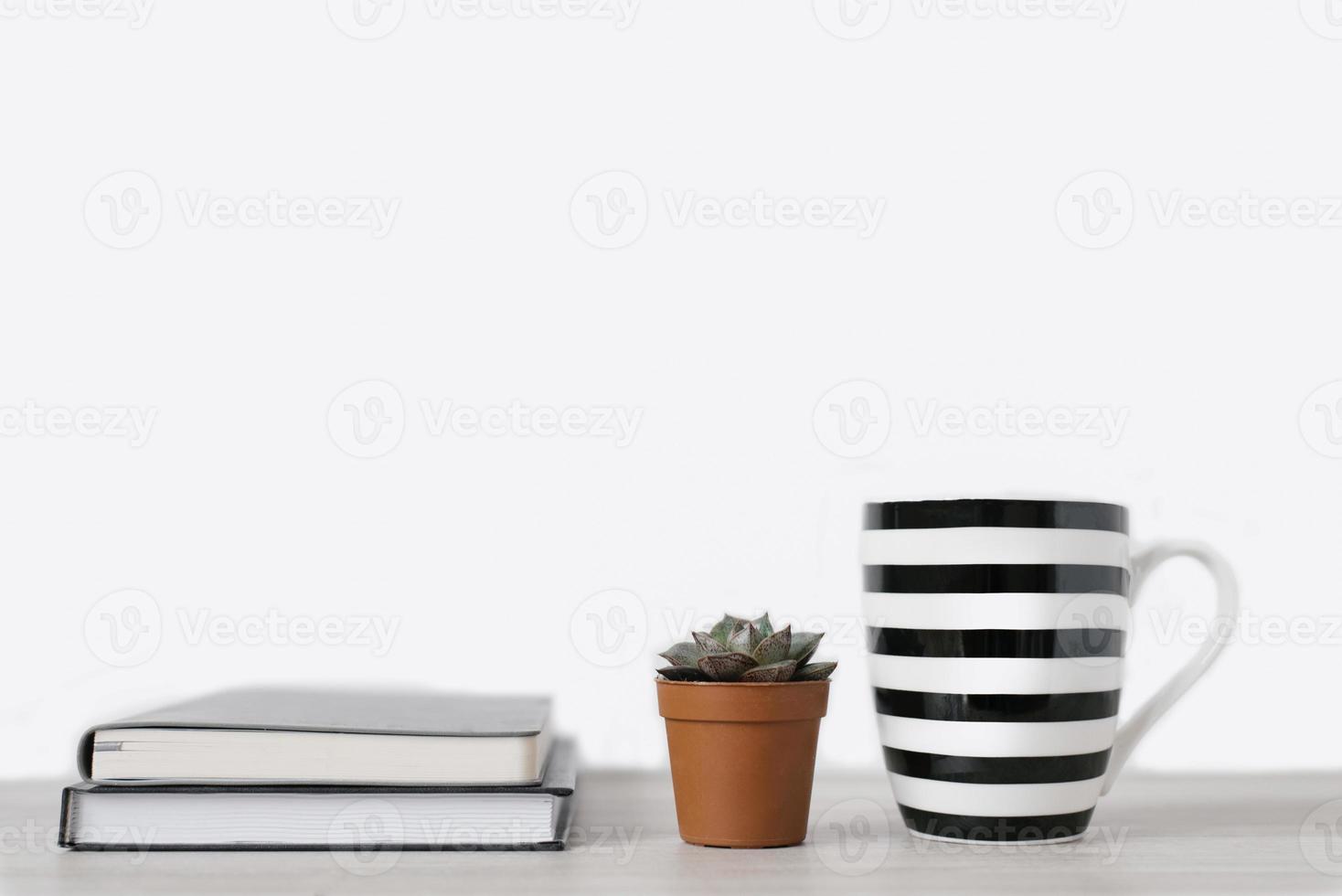 A mug with white and black stripes and notepads and a small pot of succulent with a pot of spoons on the office table. Copy space, front view photo