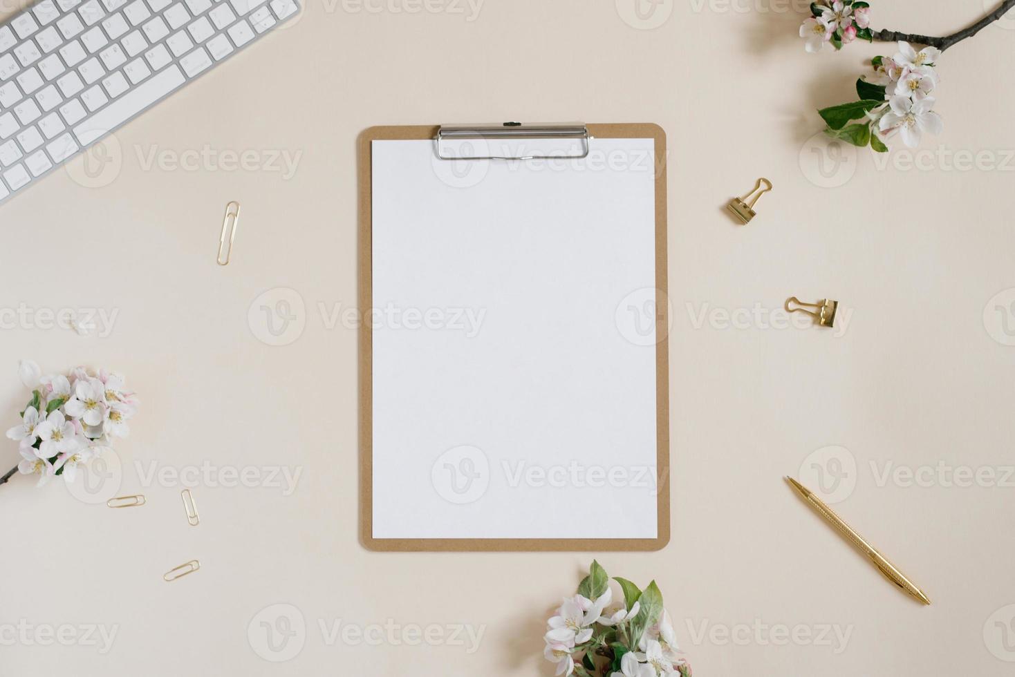 Clipboard tablet pad with blank copy space and white apple flowers, keyboard, pen and paper clips on a beige backdrop. Flat lay, top view template. Female office desk photo