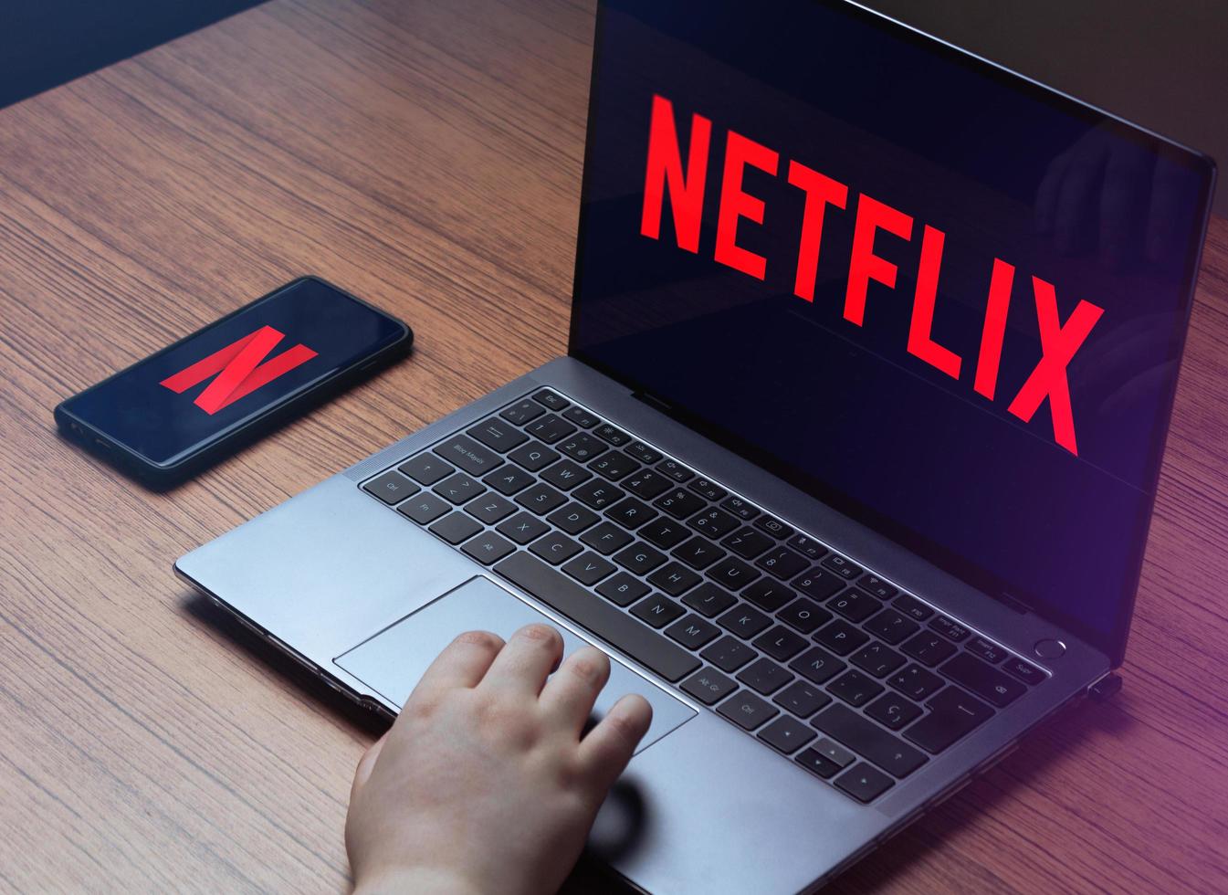 MALAGA, SPAIN, MAY 31, 2022, Young woman watching Netflix on laptop. Netflix logo on laptop and smartphone screens. Wood desk with laptop and smartphone streaming Netflix. photo