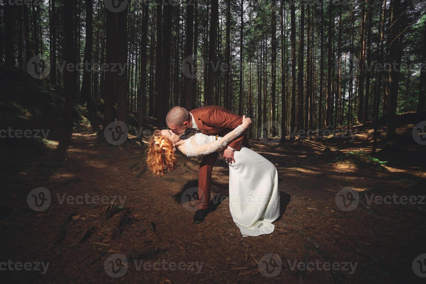 Happy stylish couple newlyweds in the green forest on summer day. bride in long white dress and groom in red suit are hugging. wedding day. photo