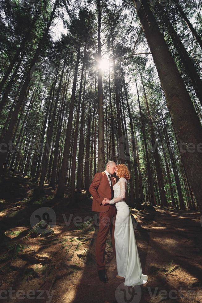 Happy stylish couple newlyweds in the green forest on summer day. bride in long white dress and groom in red suit are hugging. wedding day. photo