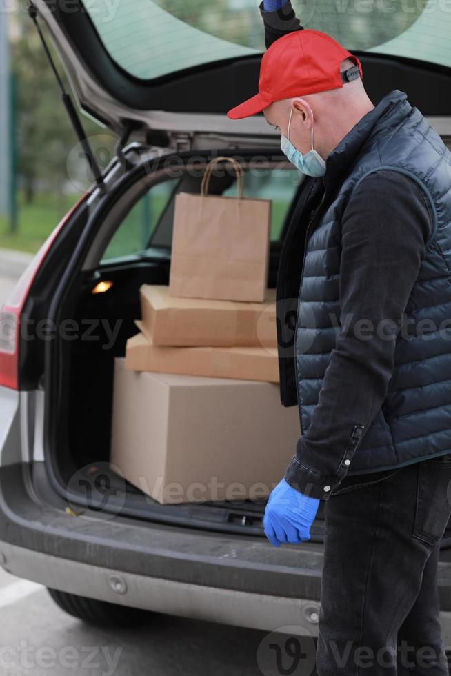 joven repartidor con máscara protectora, gorra roja y guantes cerca del camión con cajas y paquetes, al aire libre. coronavirus de servicio. las compras en línea. Bosquejo. foto