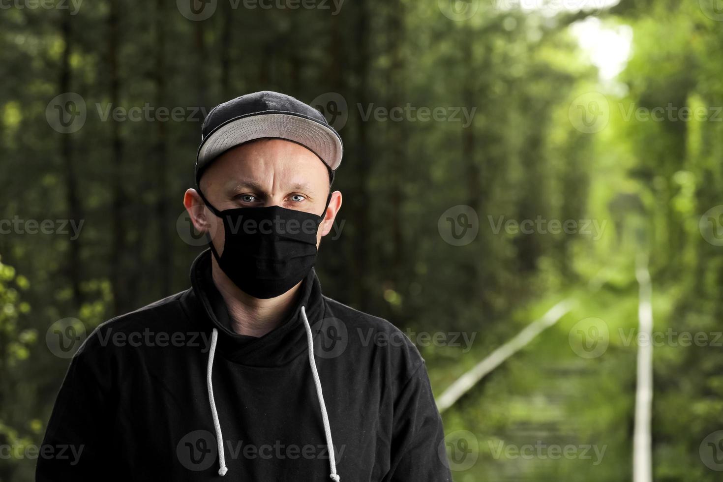 Young man in black protective antiviral mask in the summer park. The guy is resting outdoors on a sunny summer day in a mask made by his own hands. Virus protection. Quarantine measures photo