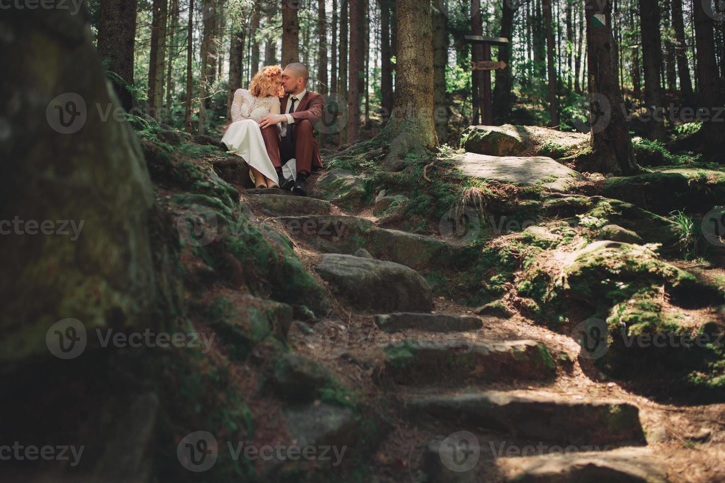 Happy stylish couple newlyweds in the green forest on summer day. bride in long white dress and groom in red suit are hugging. wedding day. photo