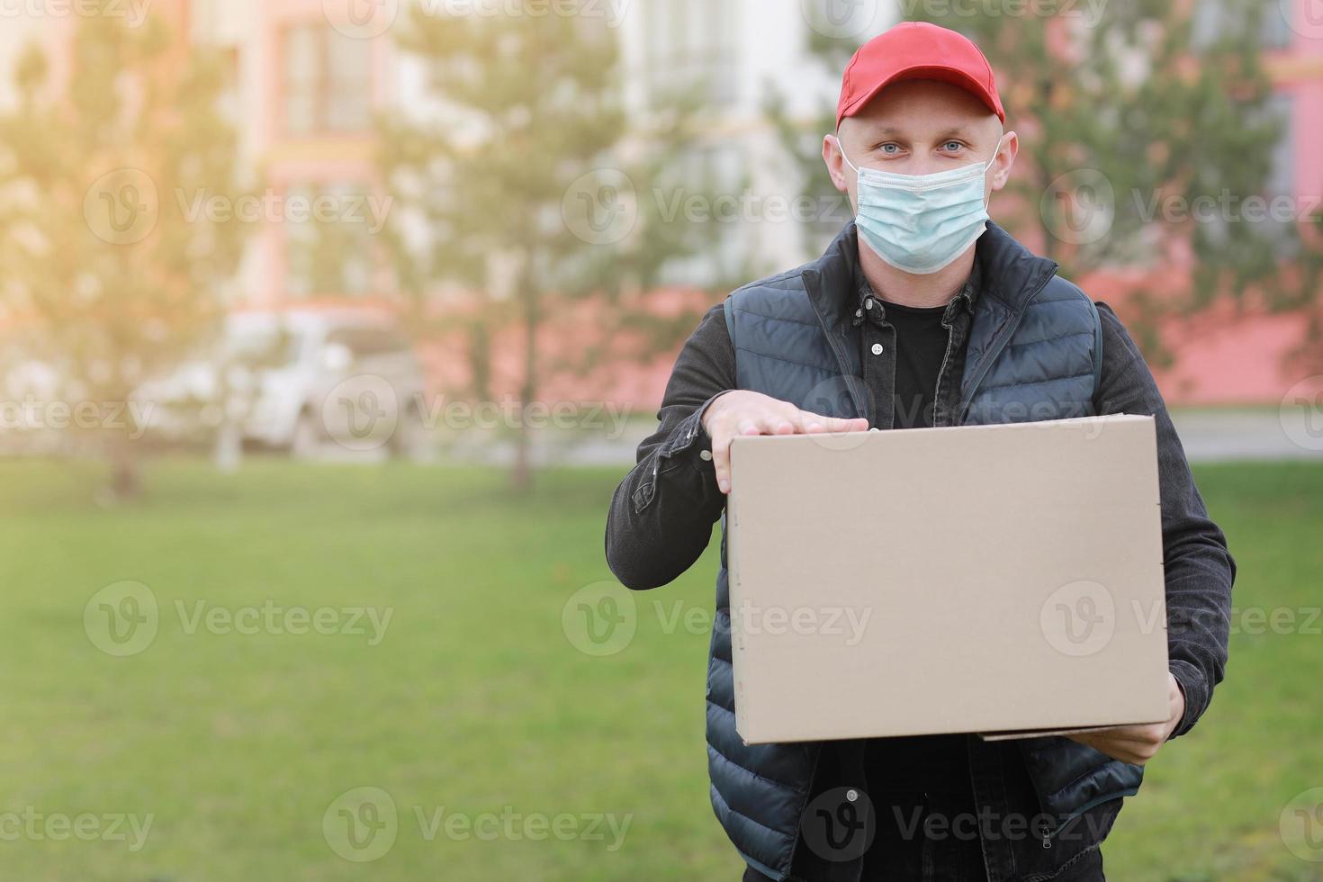 el repartidor con gorra roja, máscara médica facial sostiene una caja de cartón vacía al aire libre. coronavirus de servicio. las compras en línea. Bosquejo. foto