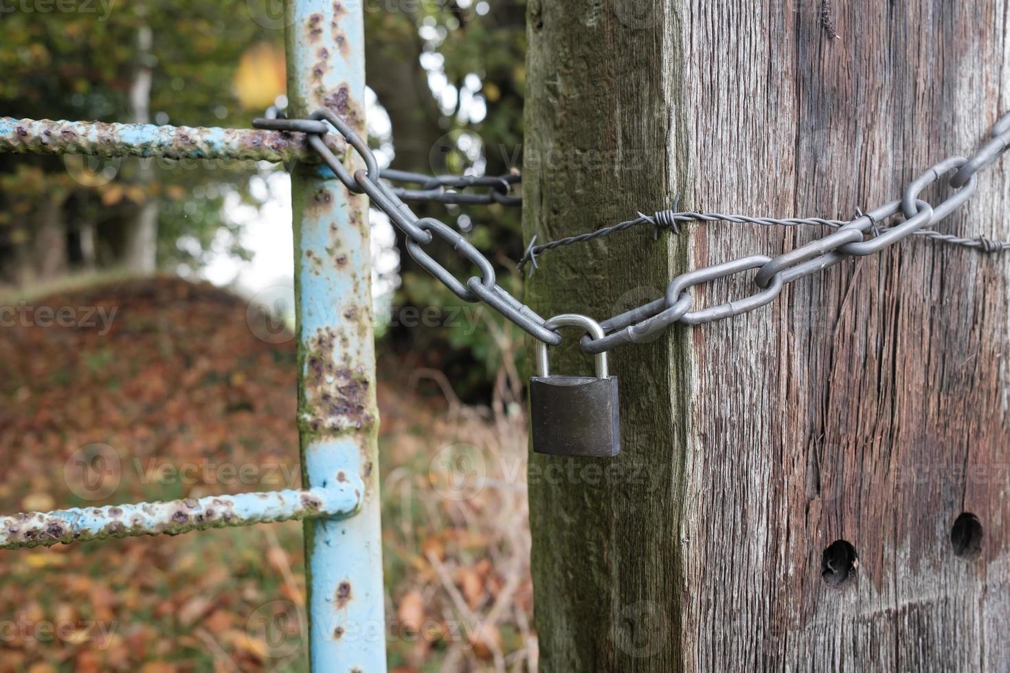 Padlock and chain linking an old metal gate and a wooden pole with barbed wire, blocking the passage to a private area. photo