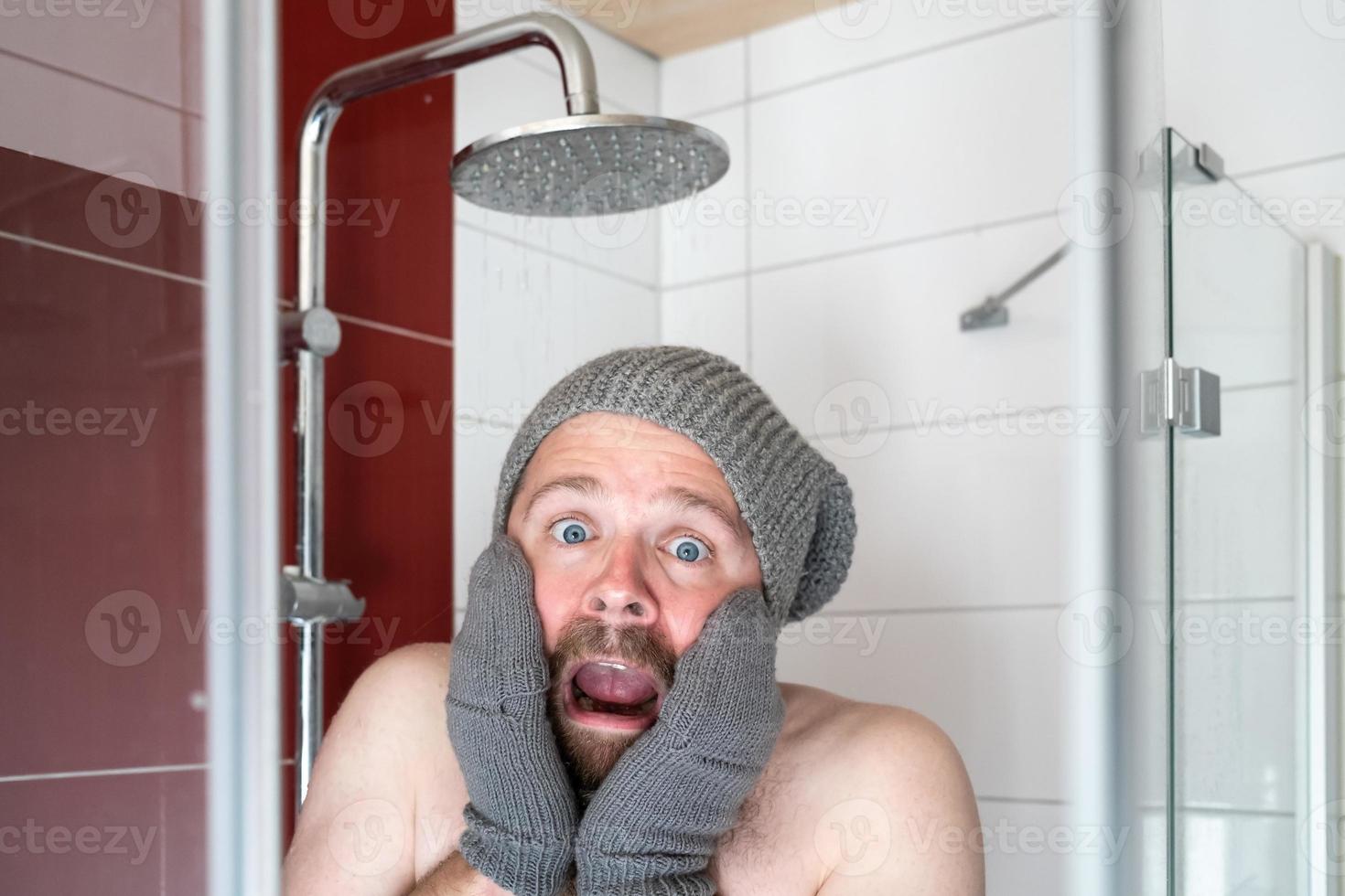 Freezing man in a hat and mittens in the shower under cold water. Concept of energy crisis. photo