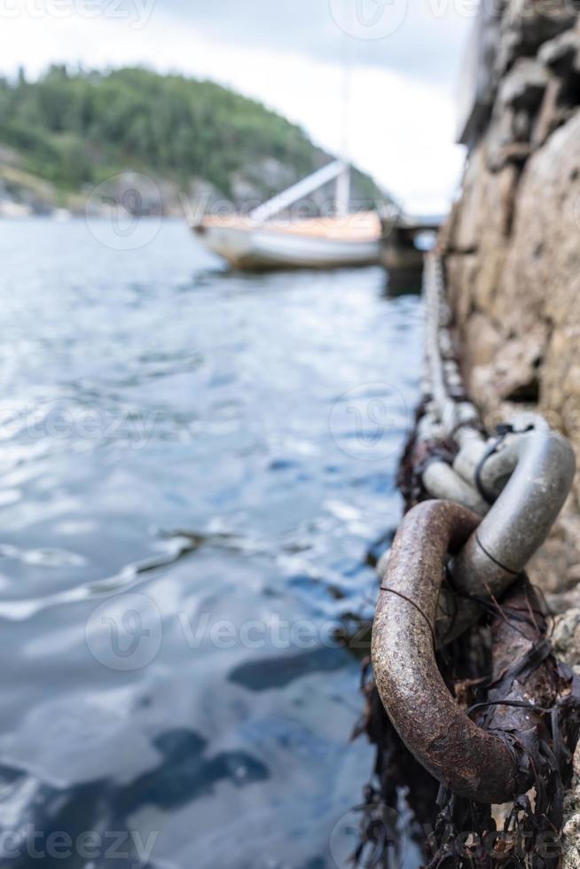 Iron chain on old stone pier, sea waves and moored sailboat and rocky shore out of focus. Bottom view. photo