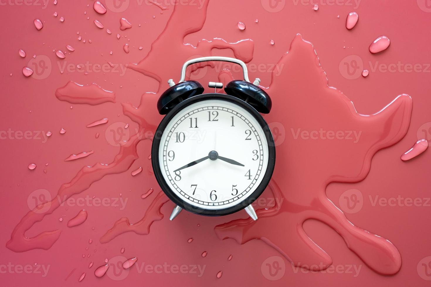 Black retro alarm clock in a pool of spilled water on a red background. Top view. photo