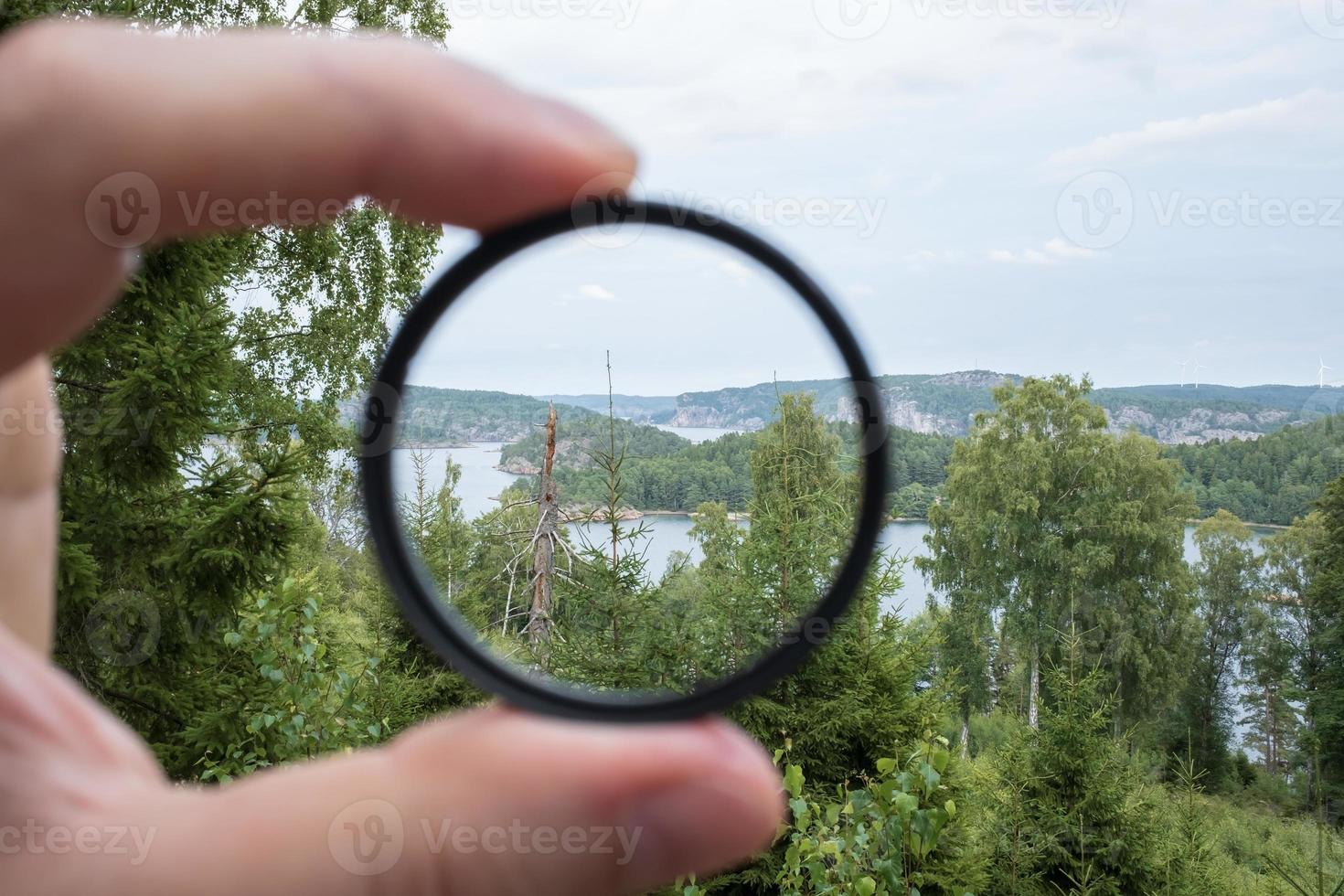 Photographer hand holds a polarizing filter, against the backdrop of trees, sky and sea. photo