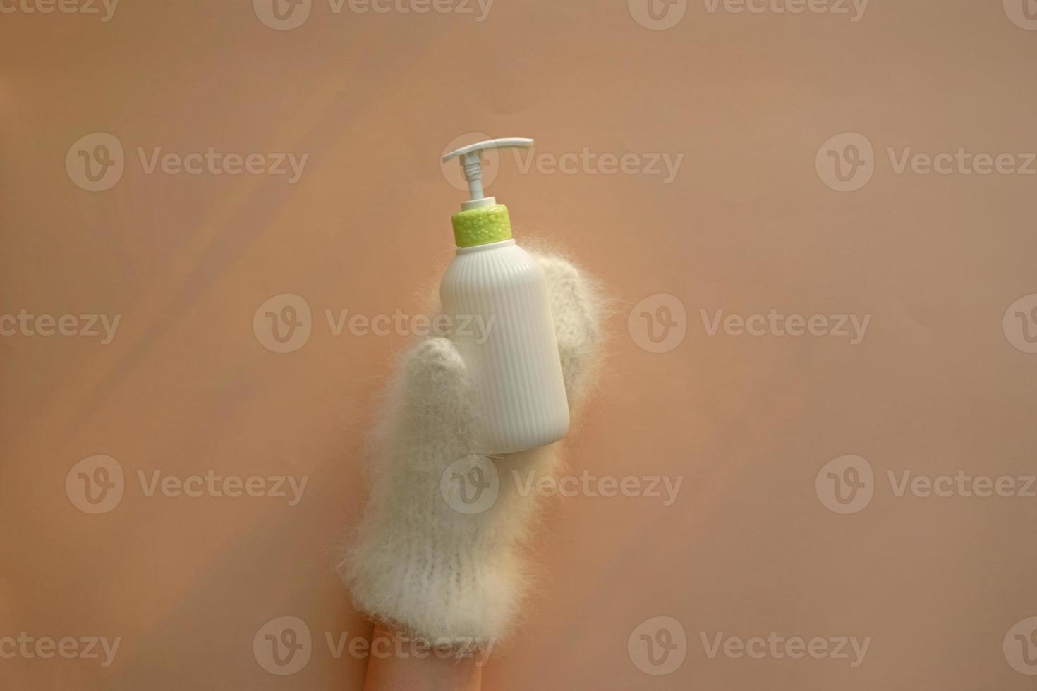 Cosmetic pump bottle of moisturizing lotion hold hands in mittens on pastel background. White corrugated bottle surface soap dispenser pump. Hand skin care lotion in winter. photo