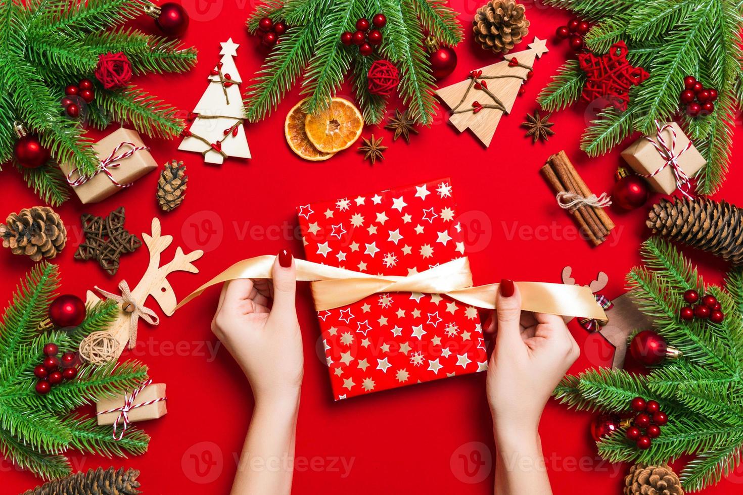 Top view of female hands holding a Christmas present on festive red background. Fir tree and holiday decorations. New Year holiday concept photo