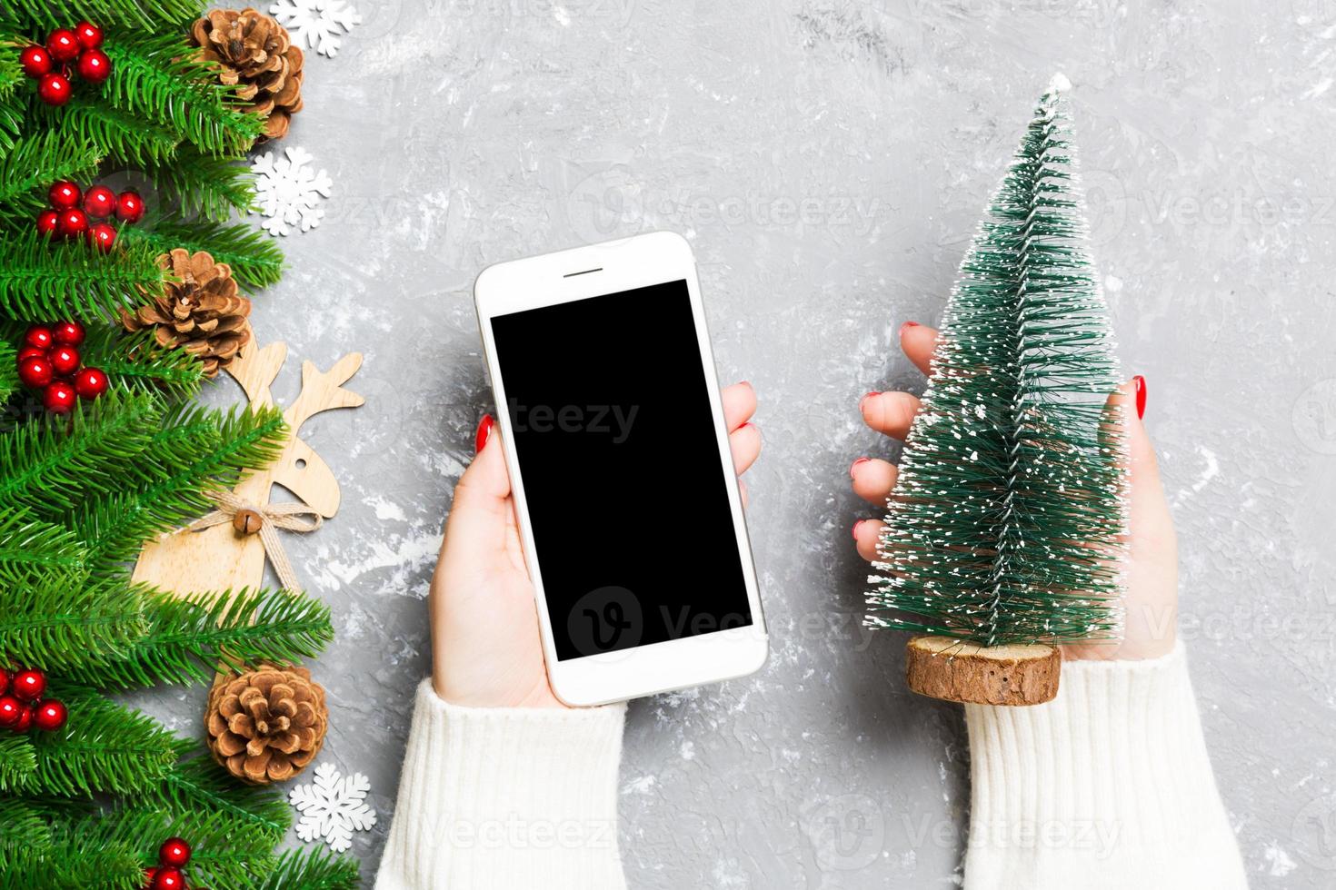 Top view of a woman holding a phone in her hand on cement New Year background made of fir tree and festive decorations. Christmas holiday concept. Mockup photo