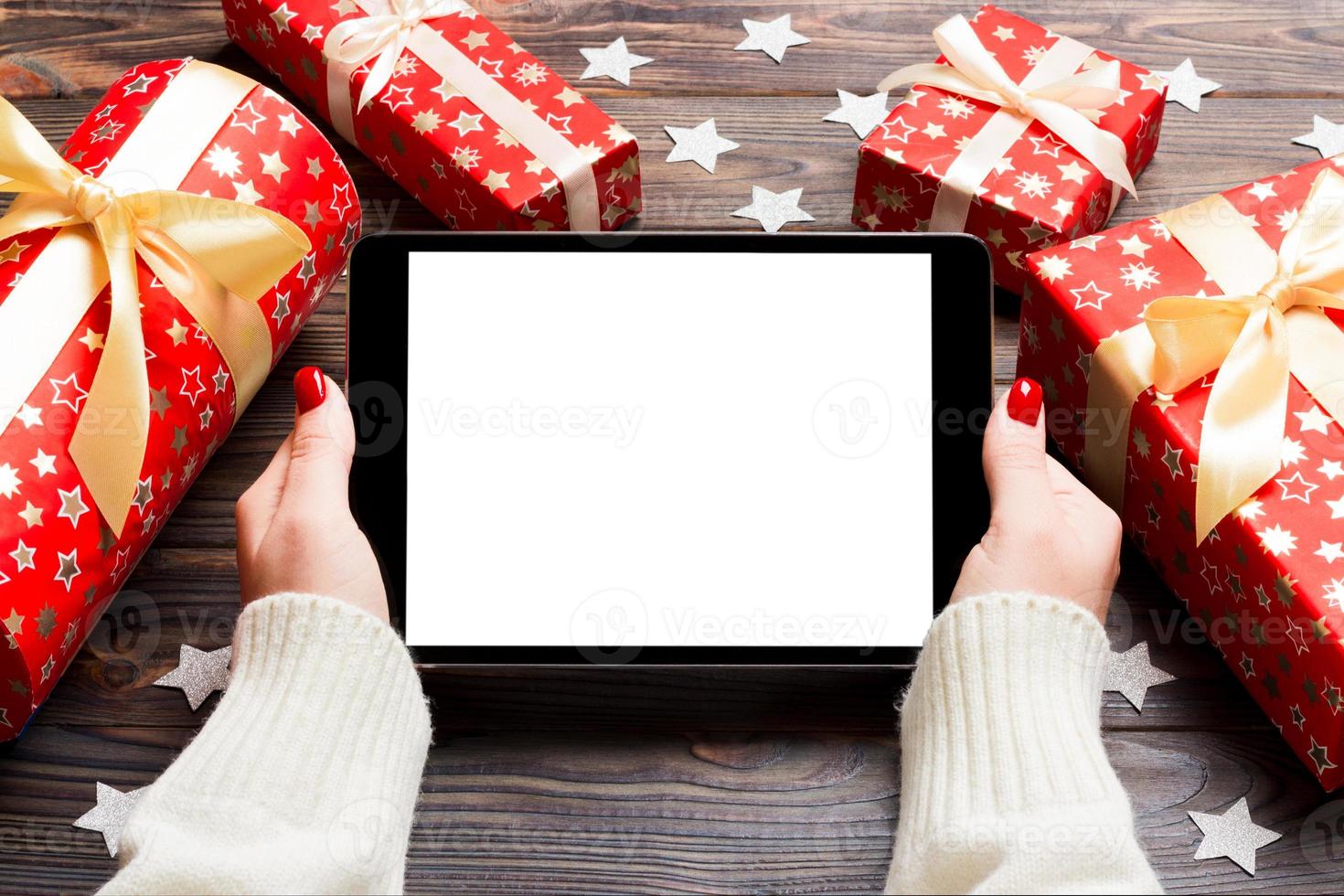 Top view of female hands holding a tablet on wooden Christmas background made of gift boxes and snowflakes. New year holiday concept. Mockup photo