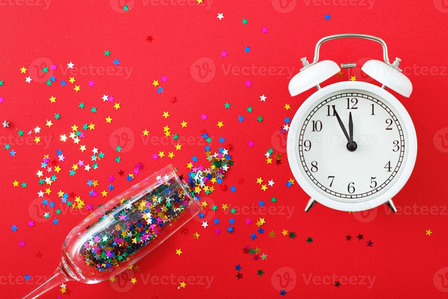 A glass with multicolored sequins, alarm clock and star shaped confettie on the red background. Party, new year, concept photo