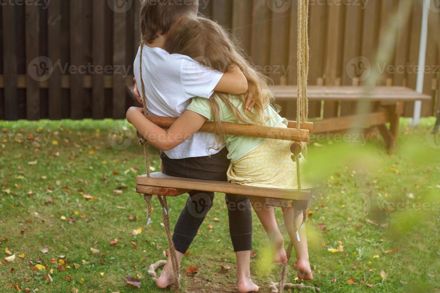 children sitting on a swing in the garden. older brother hugging little sister photo