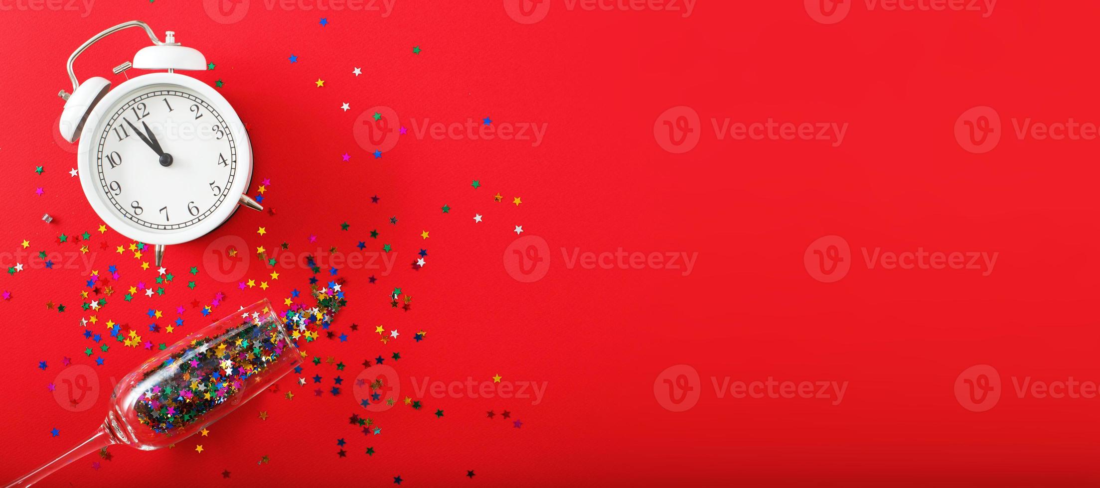 A glass with multicolored sequins, alarm clock and star shaped confettie on the red background. Banner photo