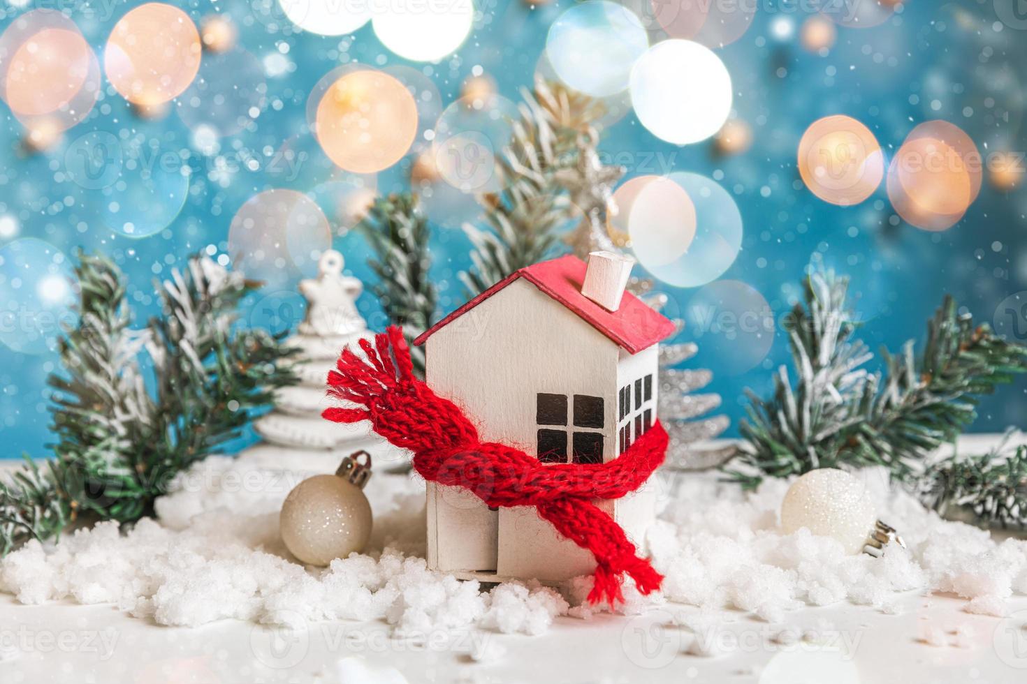 Abstract Advent Christmas Background. Toy house with knitted scarf and winter decorations ornaments on blue background with snow and defocused garland lights. Christmas with family at home concept. photo